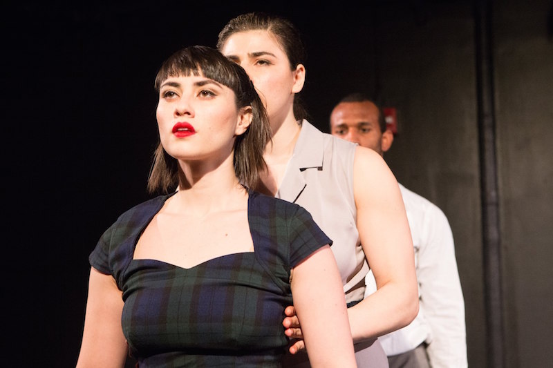 A close of a woman with red lips and bangs. Two performers stand behind her.